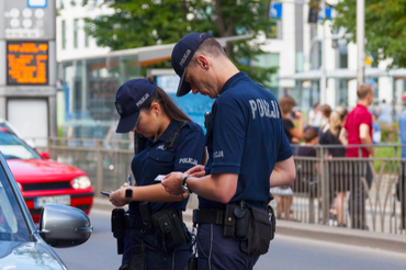 Dwoje policjantów na ulicy, sprawdzają dokumenty kierowcy w samochodzie. W tle przechodnie.