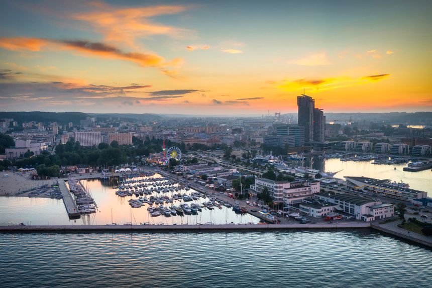 Widok na miasto przy zachodzie słońca, port z jachtami i koło młyńskie w tle.