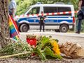 Na zdjęciu leżą kwiaty i znicz przy policyjnej furgonetce, w tle widać ludzi i tęczową flagę.