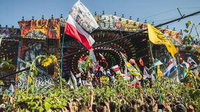 Tłum ludzi z flagami na festiwalu muzycznym, w tle kolorowa scena i błękitne niebo.