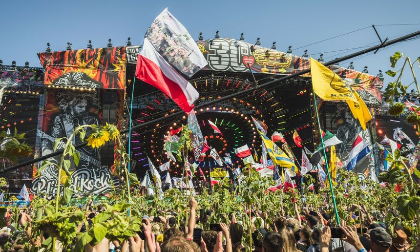 Tłum ludzi z flagami na festiwalu muzycznym, w tle kolorowa scena i błękitne niebo.