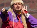 Mężczyzna z długimi blond włosami, w kolorowej kurtce, trzyma mikrofon i pozdrawia publiczność.