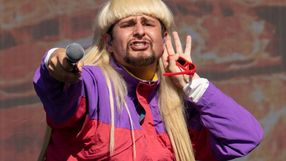 Mężczyzna z długimi blond włosami, w kolorowej kurtce, trzyma mikrofon i pozdrawia publiczność.