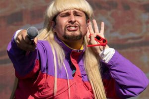 Mężczyzna z długimi blond włosami, w kolorowej kurtce, trzyma mikrofon i pozdrawia publiczność.