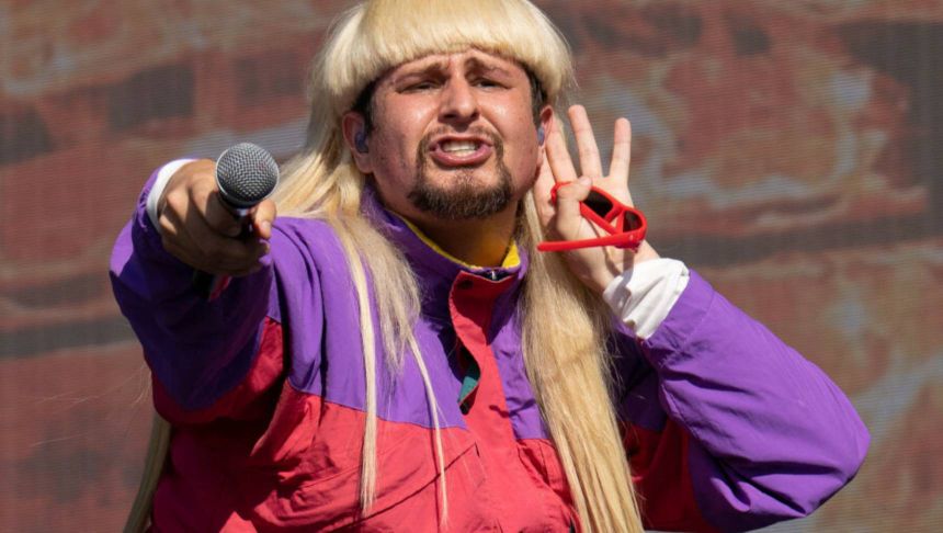 Mężczyzna z długimi blond włosami, w kolorowej kurtce, trzyma mikrofon i pozdrawia publiczność.