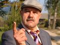 Mężczyzna w garniturze, z wąsem, wskazuje palcem, z poważnym wyrazem twarzy. Tło zielone.