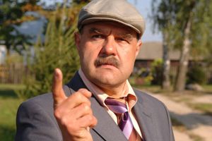 Mężczyzna w garniturze, z wąsem, wskazuje palcem, z poważnym wyrazem twarzy. Tło zielone.