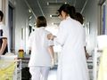 Grupa osób w białych fartuchach porusza się korytarzem szpitalnym. W tle widoczne stoły robocze.