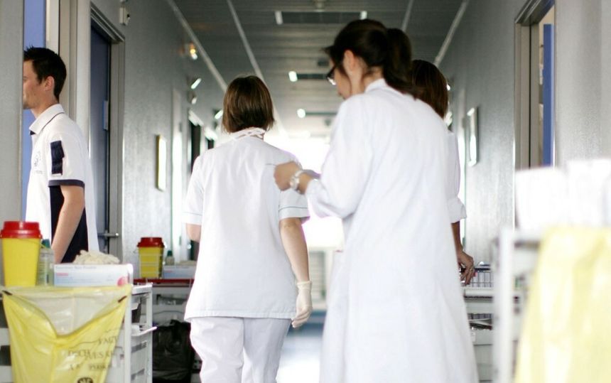 Grupa osób w białych fartuchach porusza się korytarzem szpitalnym. W tle widoczne stoły robocze.