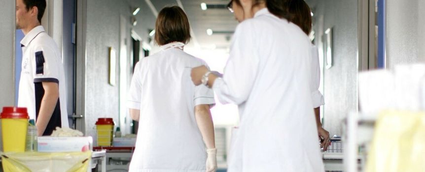 Grupa osób w białych fartuchach porusza się korytarzem szpitalnym. W tle widoczne stoły robocze.