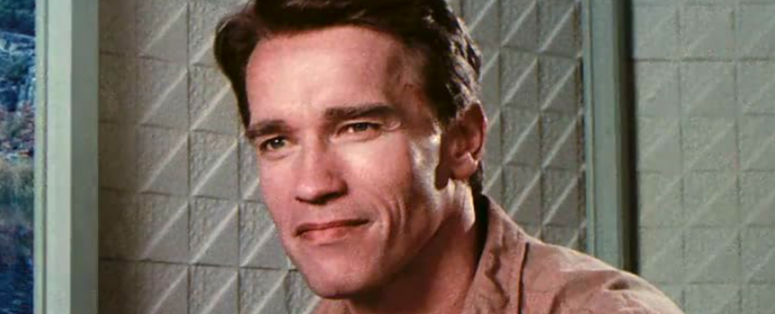 Arnold Schwarzenegger z krótkimi brązowymi włosami, uśmiecha się w jasnym otoczeniu.