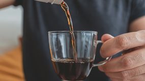 Ręka nalewa kawę do przezroczystej filiżanki na tle jasnej przestrzeni.