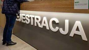 Osoba w granatowej kurtce stoi przy recepcji z napisem „REJESTRACJA”. W tle widoczny stół.