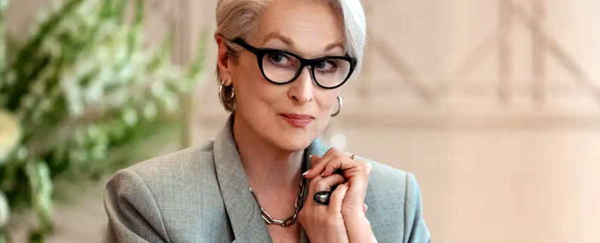 Na zdjęciu elegancka kobieta w okularach, z siwymi włosami i stylizowanym garniturem.
