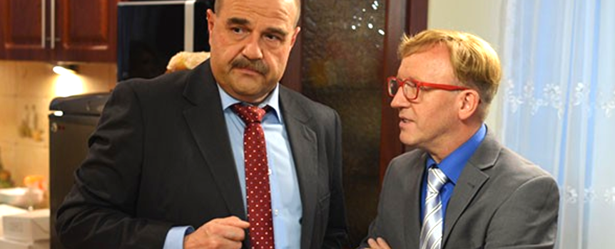 Dwaj mężczyźni w eleganckich garniturach rozmawiają w kuchni. W tle widoczna szafa i zasłony.