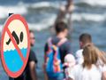Zakaz pływania, w tle plaża z ludźmi i falami. Bez rozpoznawalnych polityków.