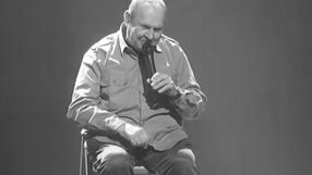 Mężczyzna w koszuli siedzi na krześle, trzyma mikrofon, śmieje się na scenie w ciemnym tle.