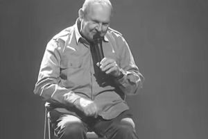 Mężczyzna w koszuli siedzi na krześle, trzyma mikrofon, śmieje się na scenie w ciemnym tle.