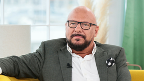 Mężczyzna w okularach, w szarym garniturze, siedzi na kanapie, wygląda na zamyślonego.