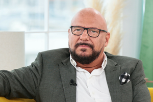 Mężczyzna w okularach, w szarym garniturze, siedzi na kanapie, wygląda na zamyślonego.