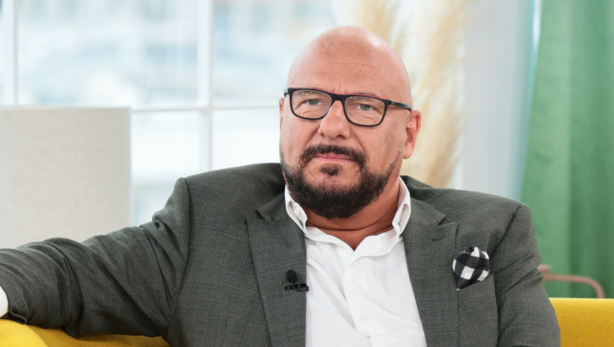 Mężczyzna w okularach, w szarym garniturze, siedzi na kanapie, wygląda na zamyślonego.