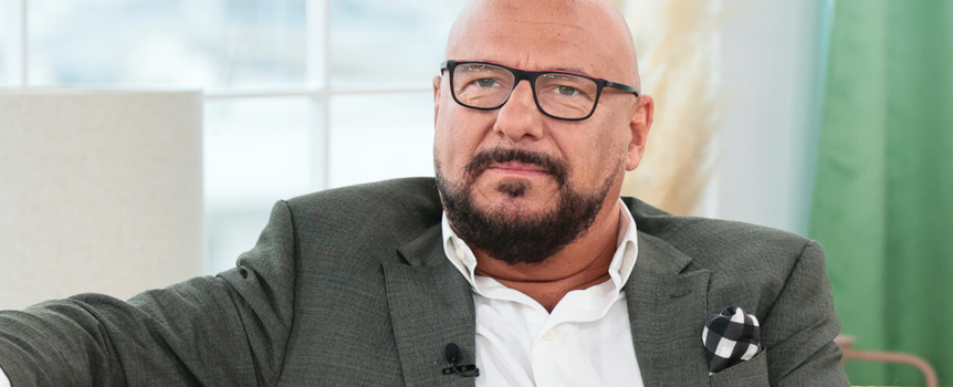 Mężczyzna w okularach, w szarym garniturze, siedzi na kanapie, wygląda na zamyślonego.