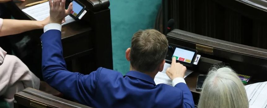 Mężczyzna w granatowym garniturze podnosi rękę w parlamencie, obok siedzi starsza kobieta.