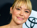 Tatiana Maslany. Uśmiechnięta osoba z jasnymi włosami i delikatnym makijażem, na tle ciemnych kolorów.