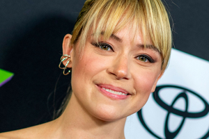 Tatiana Maslany. Uśmiechnięta osoba z jasnymi włosami i delikatnym makijażem, na tle ciemnych kolorów.