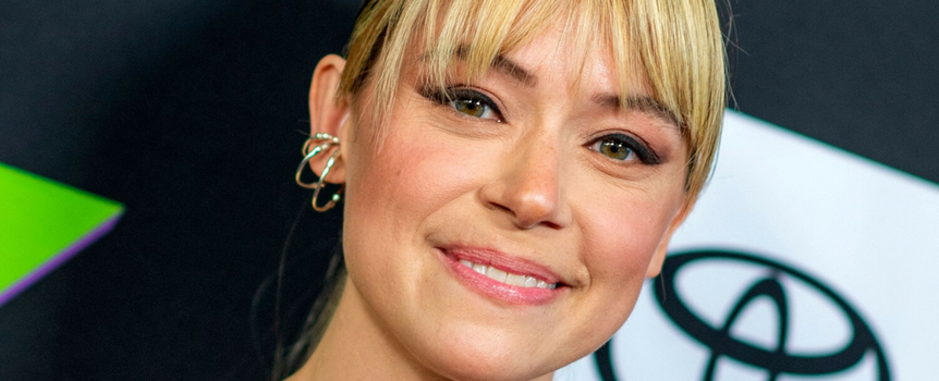 Tatiana Maslany. Uśmiechnięta osoba z jasnymi włosami i delikatnym makijażem, na tle ciemnych kolorów.