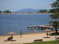 Plaża z piaskiem, wodą i drewnianym pomostem. Ludzie bawią się w wodzie i opalają się.