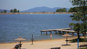 Plaża z piaskiem, wodą i drewnianym pomostem. Ludzie bawią się w wodzie i opalają się.