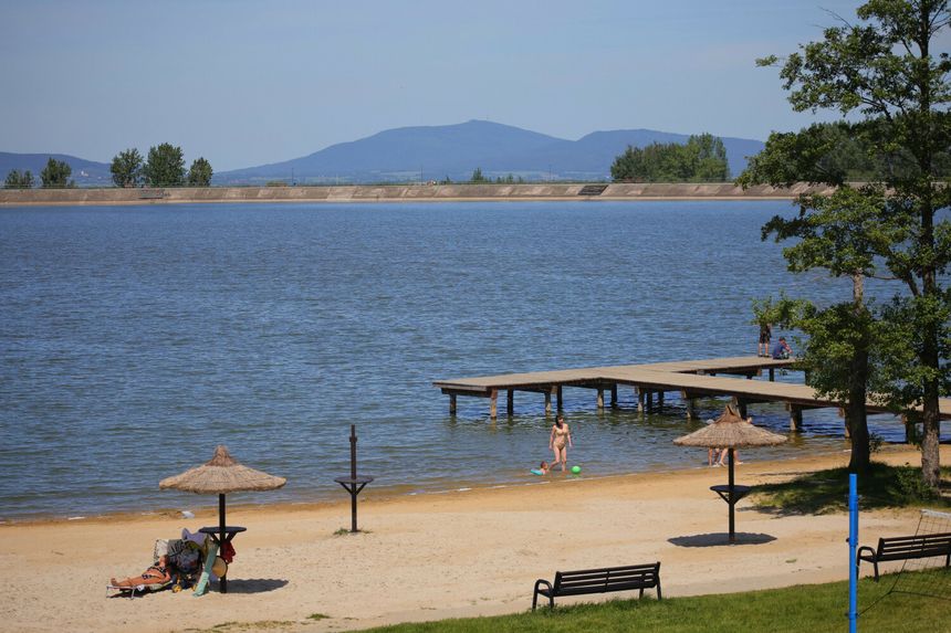Plaża z piaskiem, wodą i drewnianym pomostem. Ludzie bawią się w wodzie i opalają się.