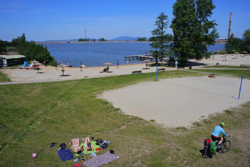 Widok na plażę z piaskiem, jezioro i osoby wypoczywające w słońcu. Rowerzysta przejeżdża obok.