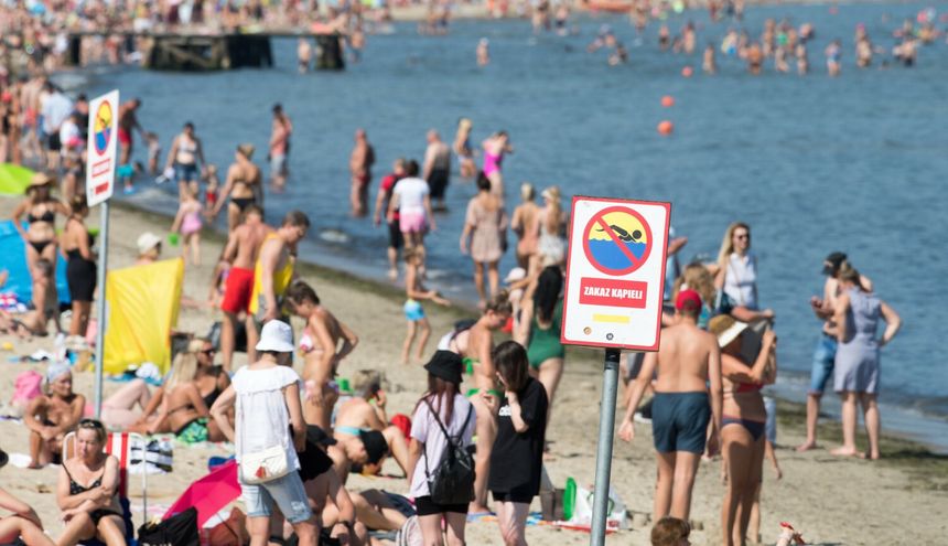 Widok plaży pełnej ludzi, znak zakazu kąpieli na pierwszym planie. Słońce, woda i wypoczywający.
