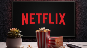 Na zdjęciu widoczna jest logo Netflixa na tle, obok popcorn i roślina doniczkowa.