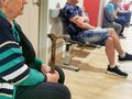 Kobieta w swetrze w paski siedzi z laską w poczekalni, wokół mężczyźni czekający na wizytę.