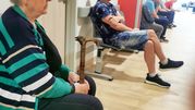 Kobieta w swetrze w paski siedzi z laską w poczekalni, wokół mężczyźni czekający na wizytę.