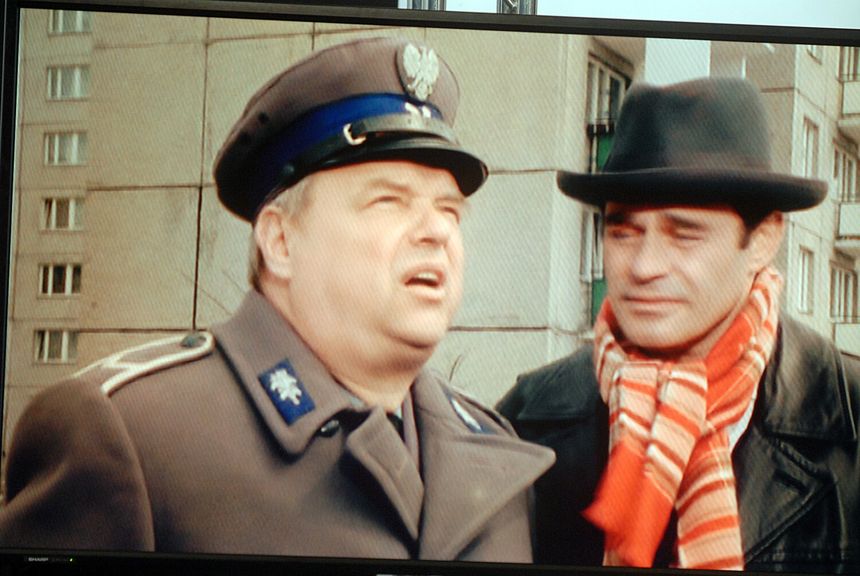 Na ekranie dwóch mężczyzn: jeden w mundurze, drugi w kapeluszu i w pasiaku. Tło z blokiem.