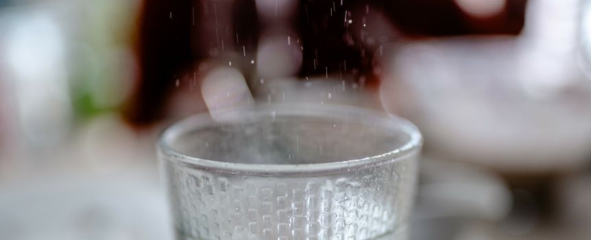 Szklanka z wodą, z której na górze spadają krople. W tle zamazane sylwetki osób.