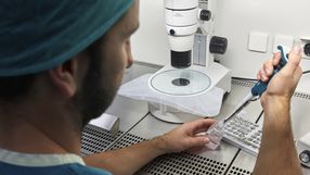 Osoba w zielonym uniformie laboratoryjnym obsługuje pipetę w laboratorium. W tle mikroskop.