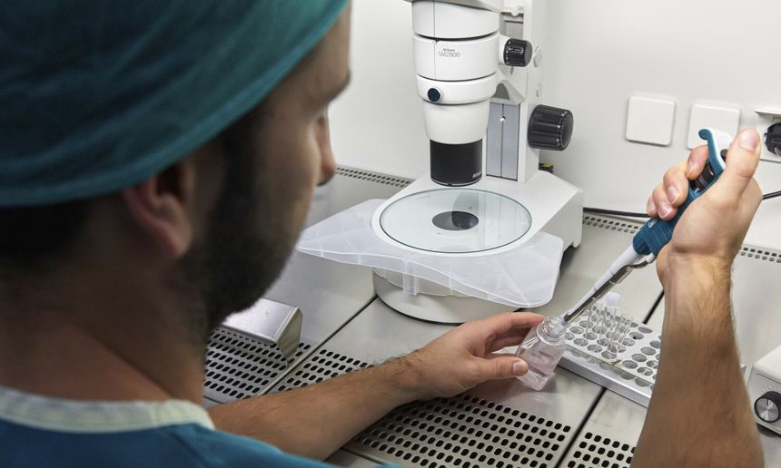 Osoba w zielonym uniformie laboratoryjnym obsługuje pipetę w laboratorium. W tle mikroskop.