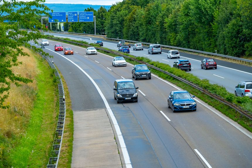 Ruch samochodowy na autostradzie, z zielonymi drzewami po bokach. Znaki drogowe w tle.