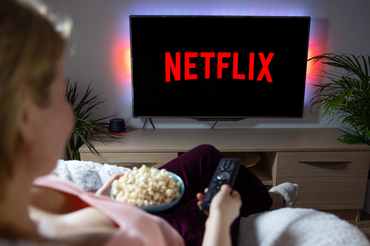 Kobieta siedzi na kanapie, trzymając miskę popcornu, patrzy na TV z napisem Netflix.