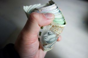 Ręka trzyma zwinięty plik banknotów, widoczny jeden z polskich banknotów z portretem.