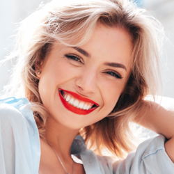 Uśmiechnięta kobieta z blond włosami w jasnej koszuli, na tle miejskiego krajobrazu.