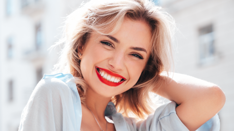 Uśmiechnięta kobieta z blond włosami w jasnej koszuli, na tle miejskiego krajobrazu.