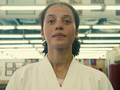 Na zdjęciu młoda kobieta w białym kimono, stojąca w sali treningowej z oświetleniem w tle.