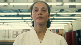 Na zdjęciu młoda kobieta w białym kimono, stojąca w sali treningowej z oświetleniem w tle.