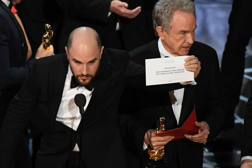 Dwóch mężczyzn na scenie Oscarów, jeden trzyma statuetkę, drugi kartkę z nazwą filmu \